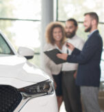 Close up of white car headlight. Happy couple standing near car dealer. discussing purchase of vehicle. Manager in dark blue jacket holding keys and black folder, showing documents.