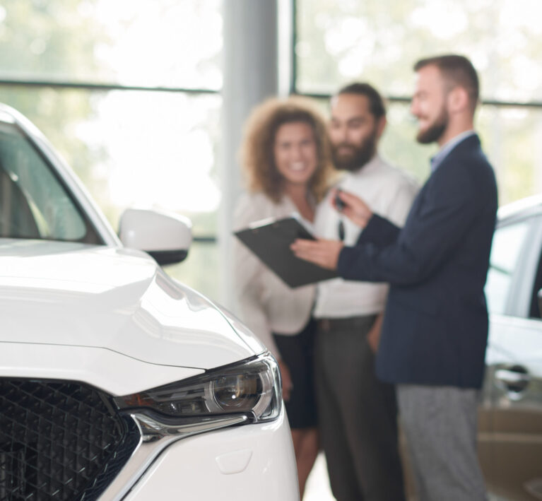 Close up of white car headlight. Happy couple standing near car dealer. discussing purchase of vehicle. Manager in dark blue jacket holding keys and black folder, showing documents.